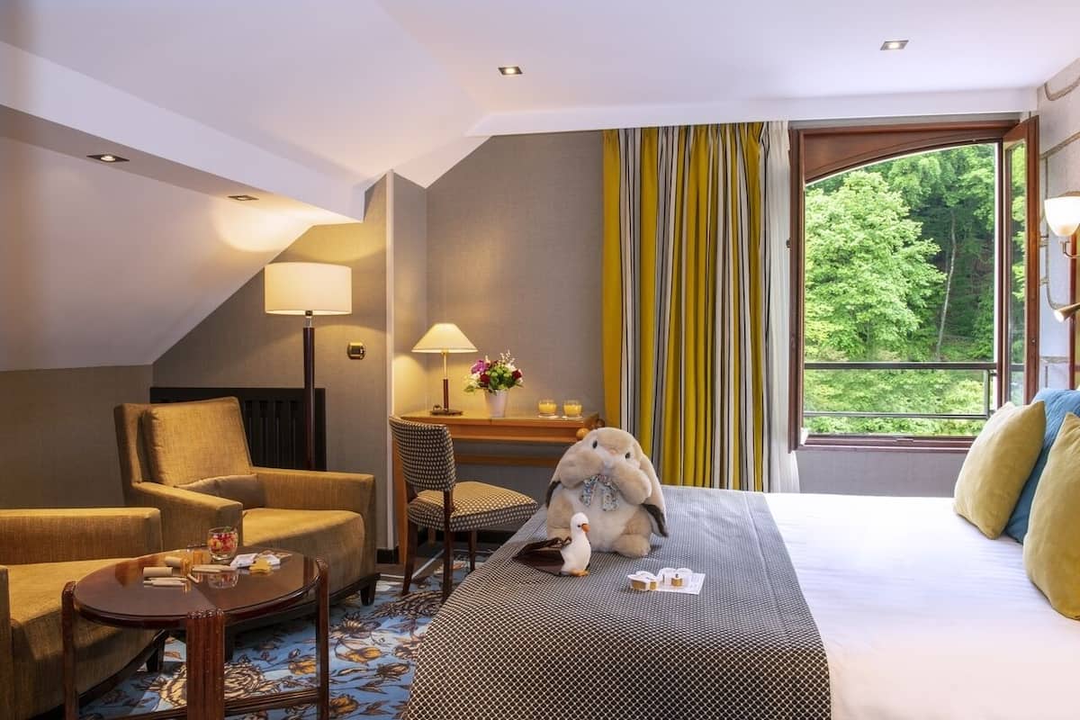Habitación de hotel con cama, dos sillones, una mesita, un escritorio, una lámpara, una ventana con cortinas y vistas al exterior con vegetación.