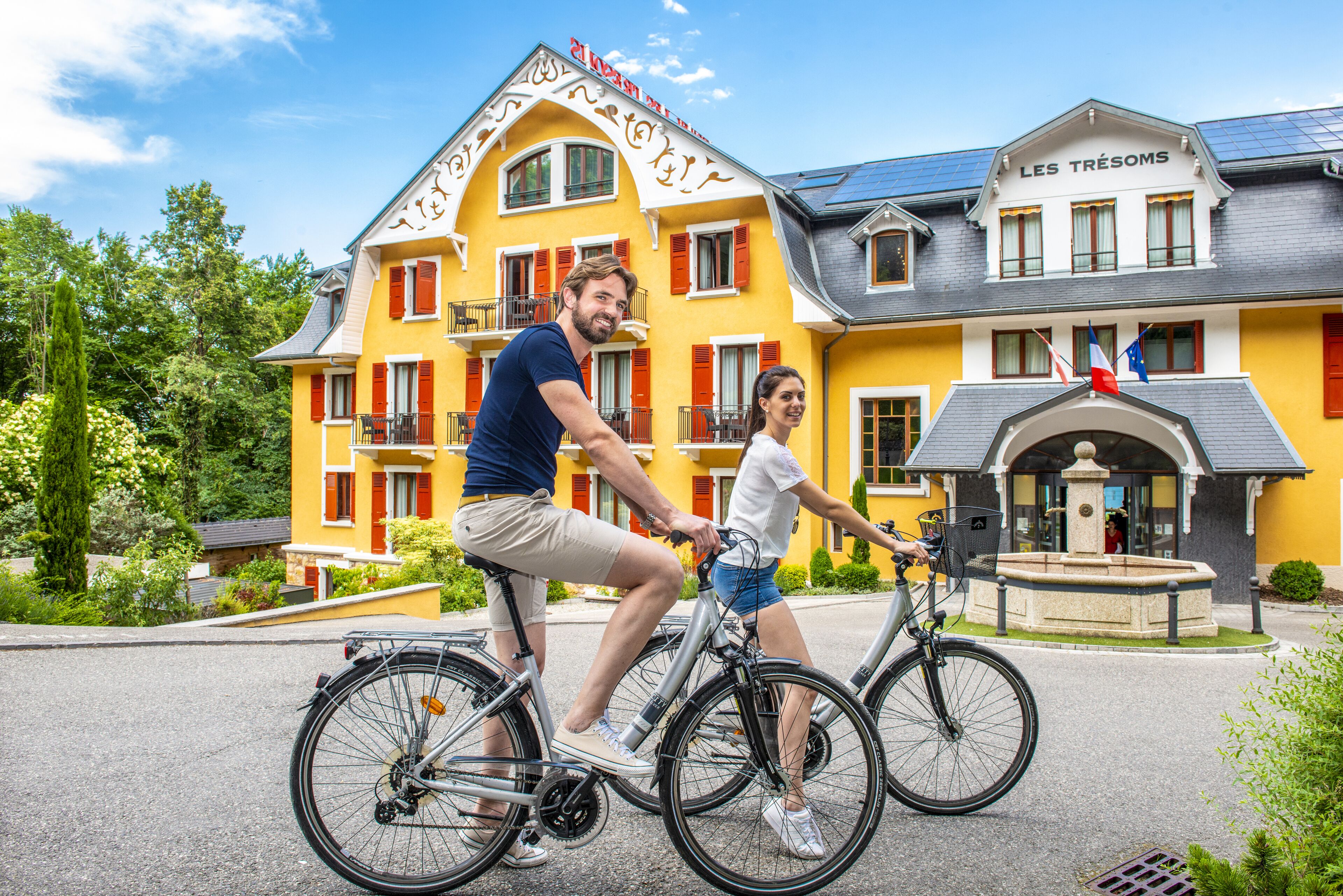 Un bâtiment jaune avec une enseigne indiquant « LES TRÉSOMS » et une fontaine devant.
