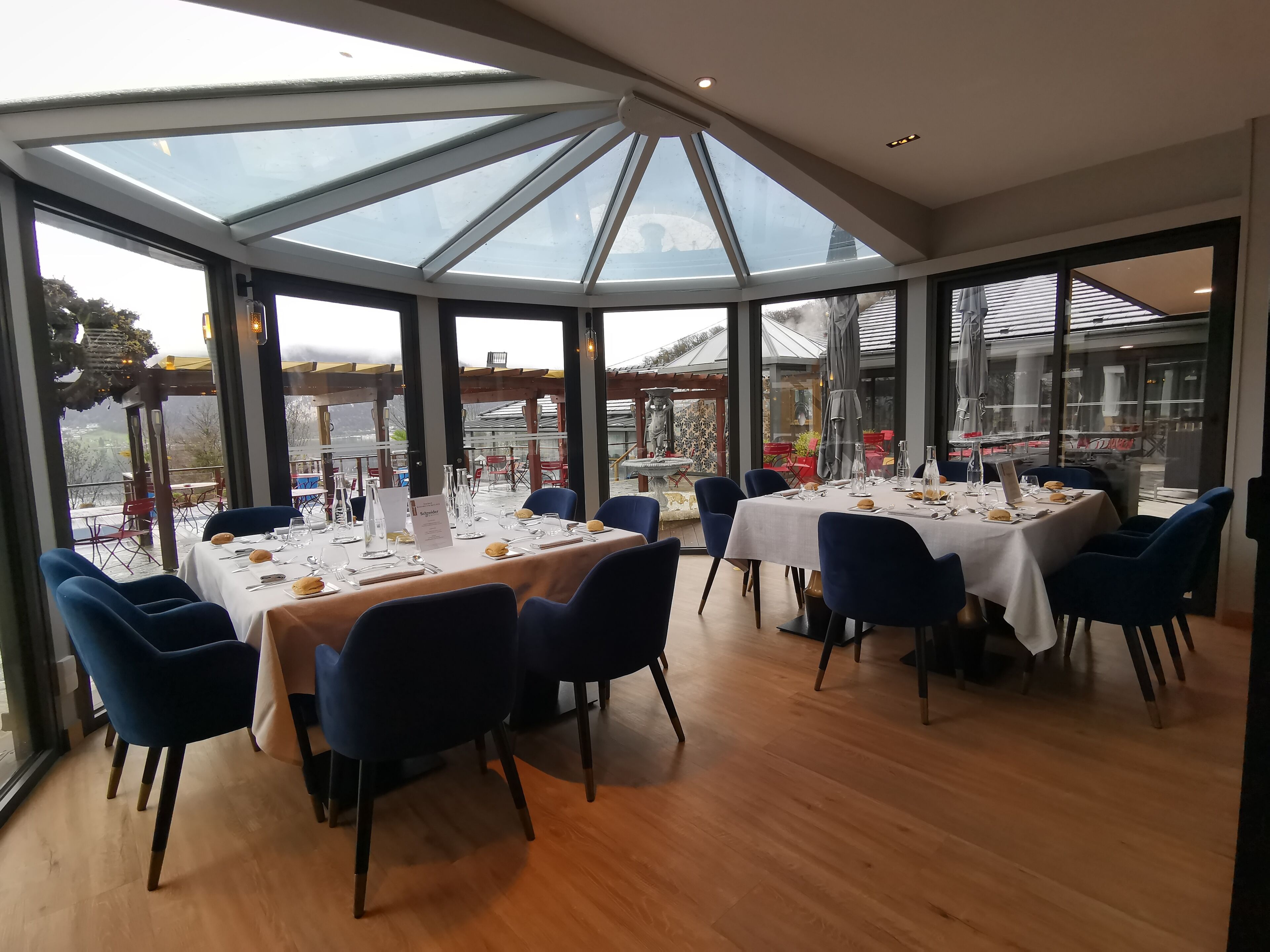 A restaurant with a glass ceiling, wooden flooring, and blue chairs. Tables are set with white tablecloths and silverware.