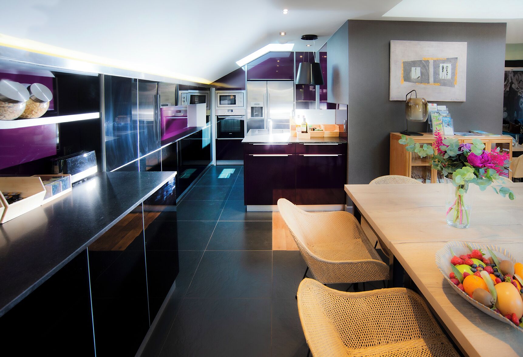 A modern kitchen with built-in appliances, a dining area with wicker chairs, and a vase with flowers on the table.