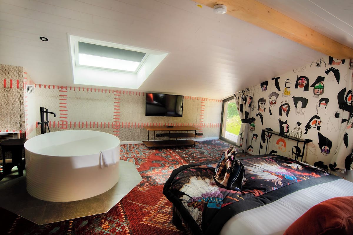 Un dormitorio con tragaluz, una cama grande con cabecera colorida, una bañera blanca y un televisor en la pared.