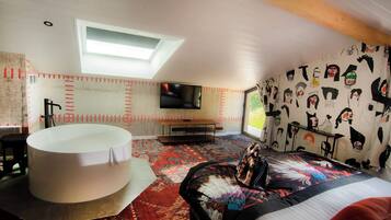 Un dormitorio con tragaluz, una cama grande con cabecera colorida, una bañera blanca y un televisor en la pared.