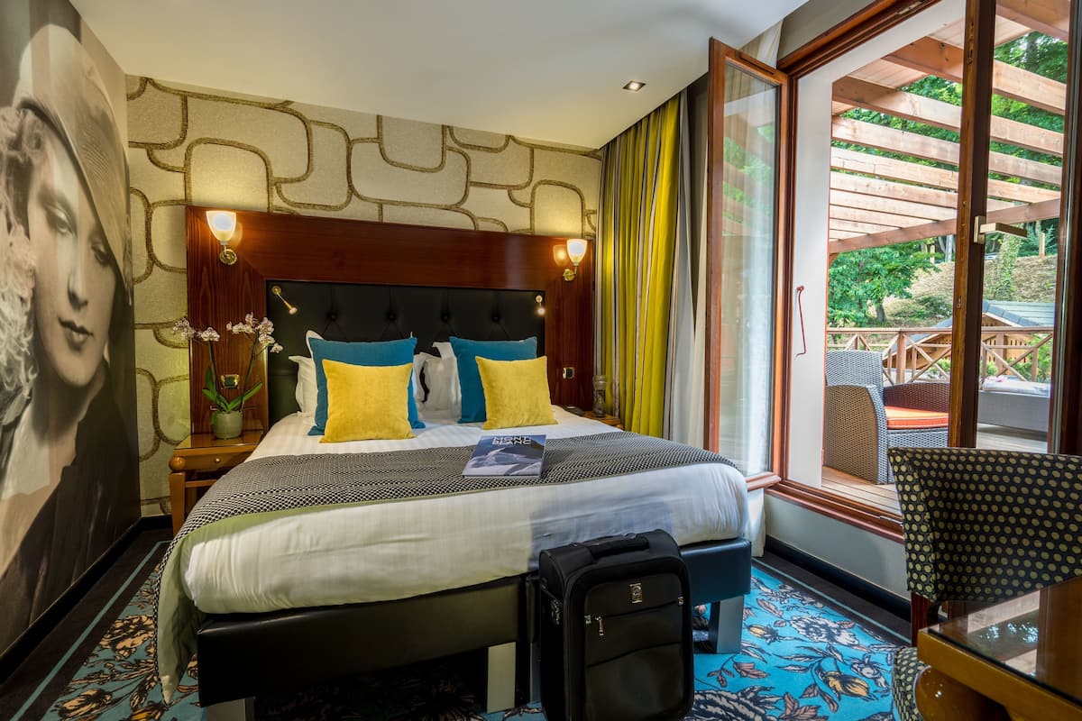 Habitación de hotel con una cama grande, cabecera con iluminación integrada, vista al exterior por una puerta de vidrio y una pequeña mesa con una silla.