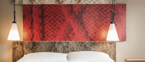 A hotel room with a bed, two pendant lights, a red and gray textured wall art, and a bedside table with a phone.
