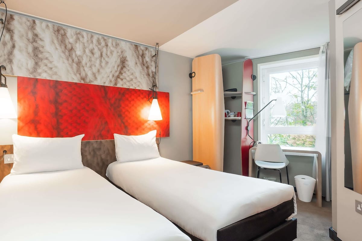 Une chambre d’hôtel avec deux lits, un bureau, une chaise et une fenêtre donnant sur des arbres.