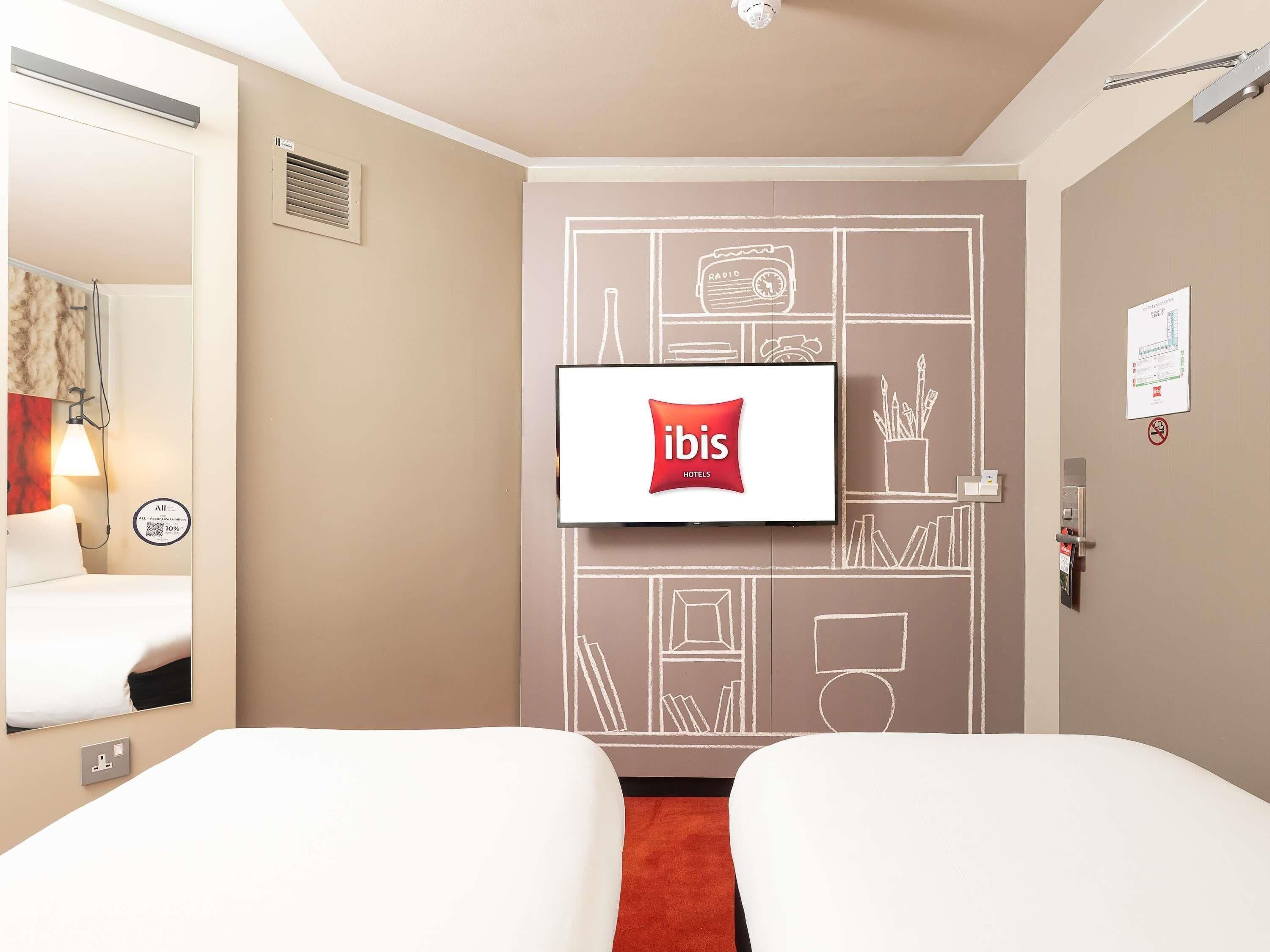 Hotellihuone, jossa on kaksi sänkyä, seinälle asennettu televisio ja valkoisella kehyksellä varustettu ovi.