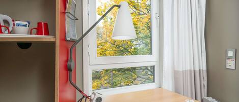 Een kamer met een bureau, een stoel, een lamp, een raam met herfstkleurige bladeren buiten en een radiator.
