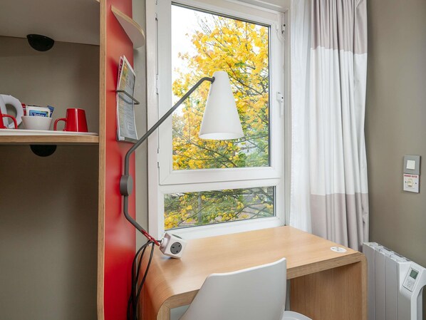 Een kamer met een bureau, een stoel, een lamp, een raam met herfstkleurige bladeren buiten en een radiator.