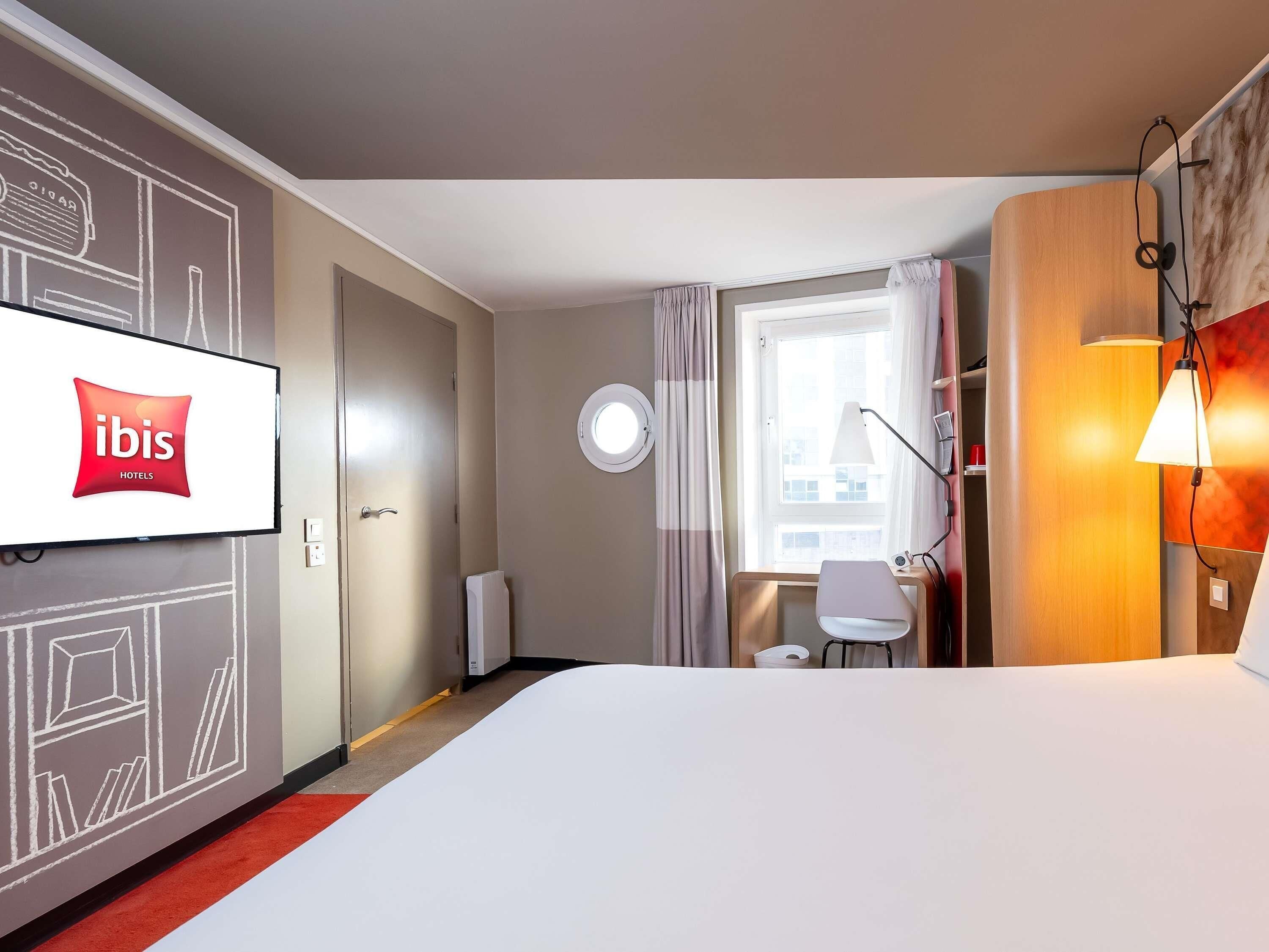 Une chambre d’hôtel comprenant un lit, un bureau, une chaise, un téléviseur affichant le logo ibis, une fenêtre avec des rideaux et une lampe.