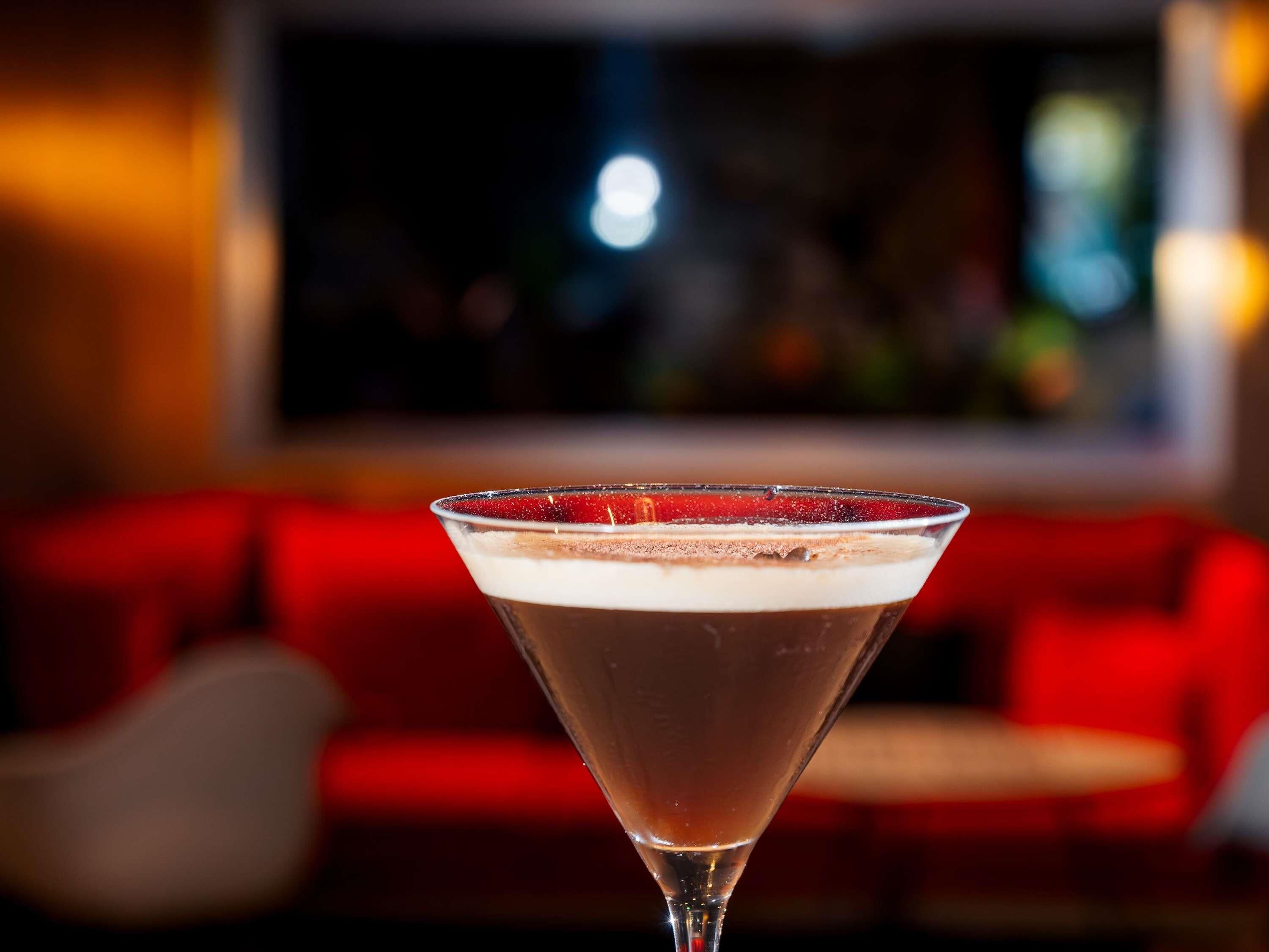 Un verre à martini, surmonté d’une mousse onctueuse, est posé sur une table dans une pièce faiblement éclairée, avec un canapé rouge en arrière-plan.