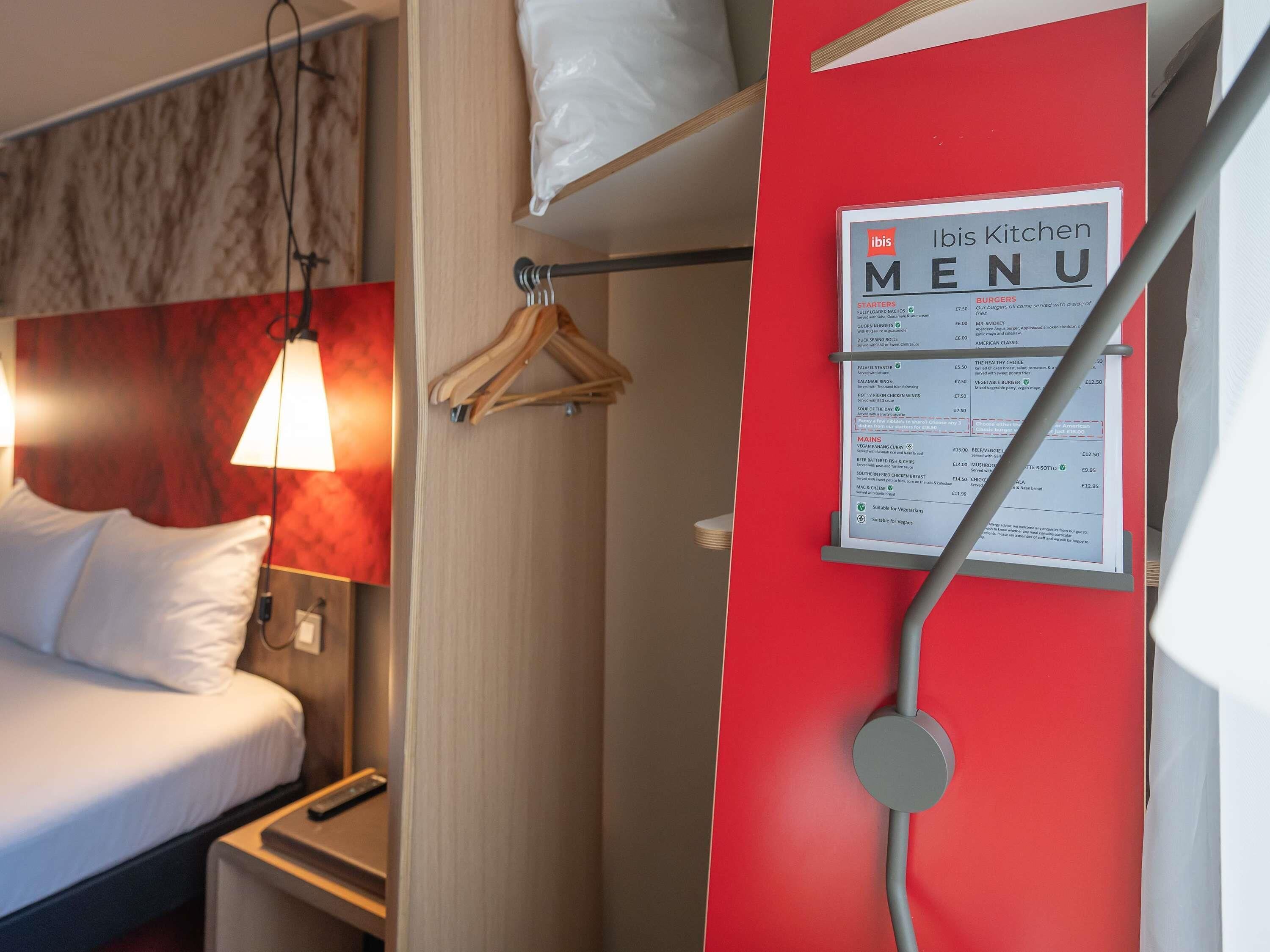 Hotellihuone, jossa on sänky, yöpöytälamppu ja punainen seinä. Huoneessa on myös minibaari ja ruokalista.