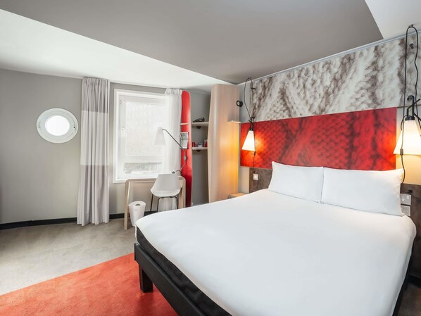 Een hotelkamer met een bed, een bureau, een stoel, een raam met gordijnen en een rode accentmuur.