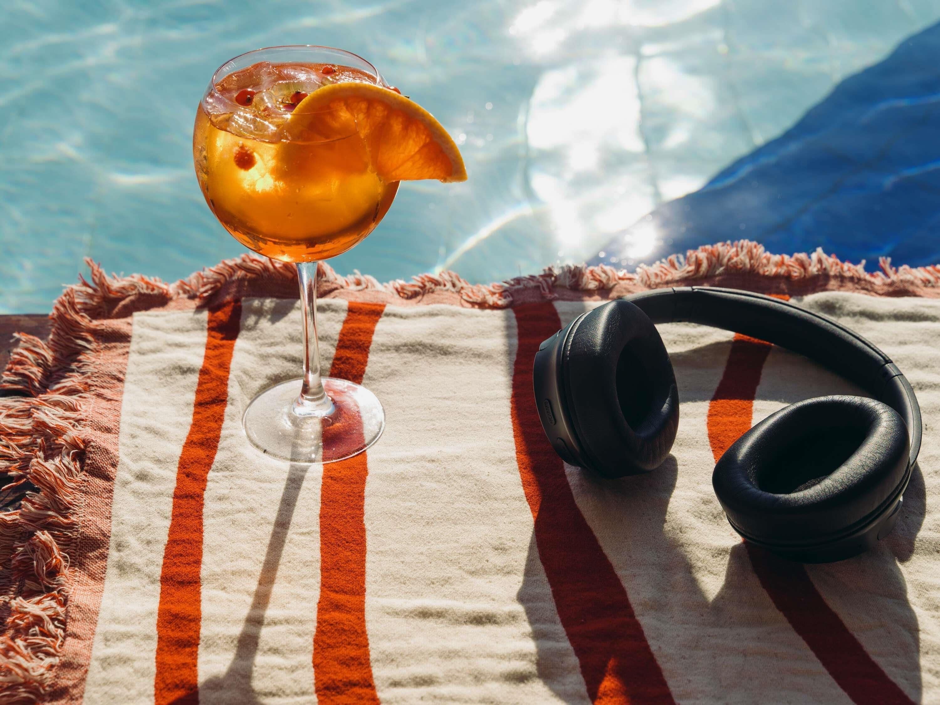A glass with a beverage, headphones, and a striped towel by a pool.