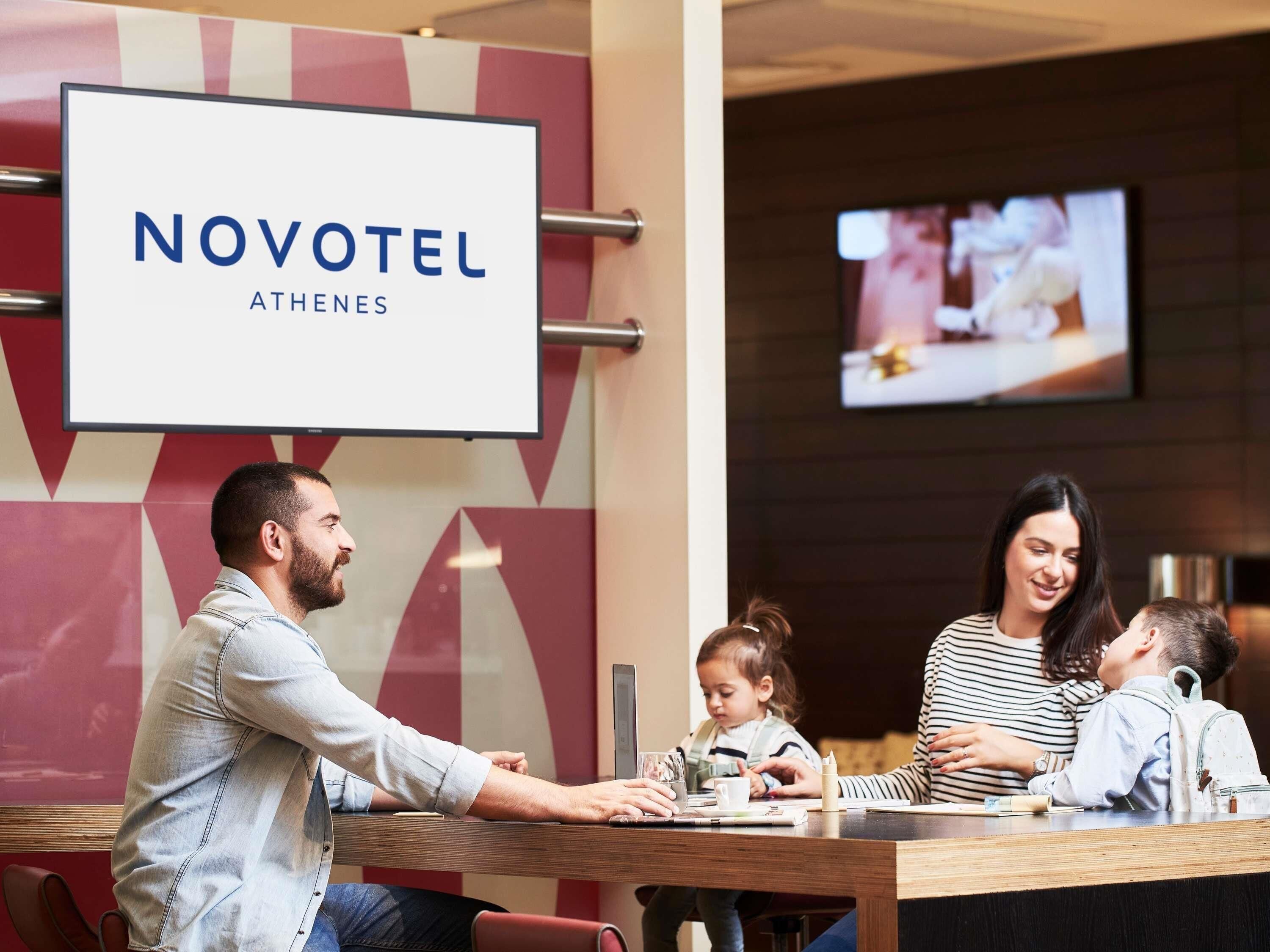 Una hall d'albergo con un grande cartello che recita 'NOVOTEL ATHENES', una reception e uno schermo televisivo sullo sfondo.