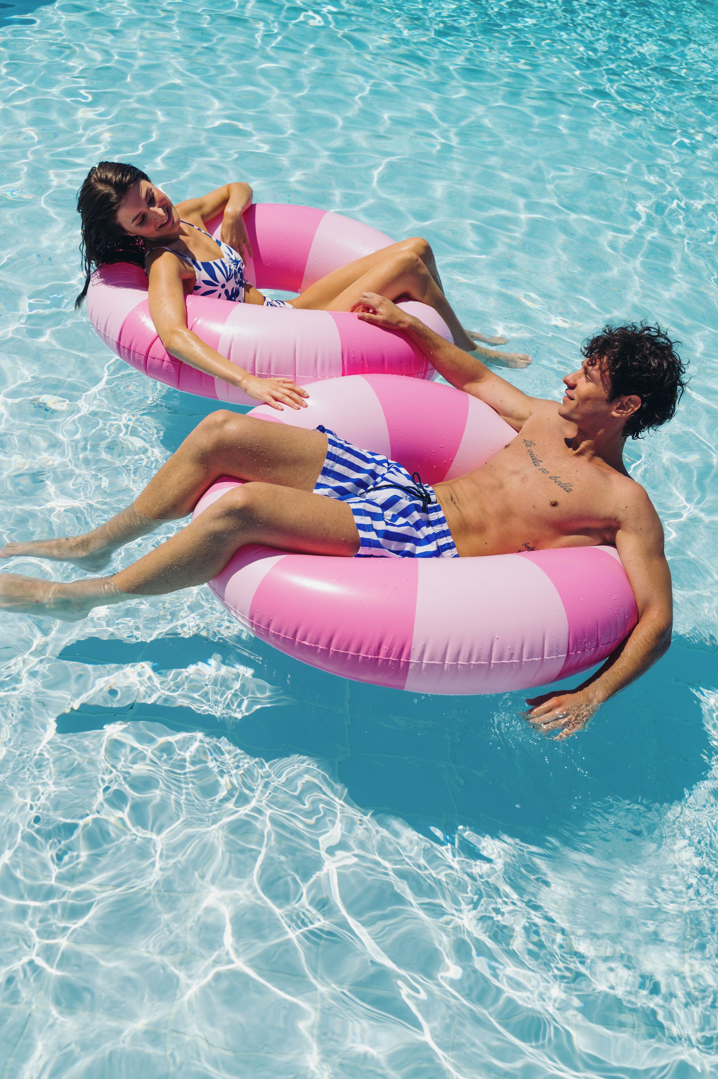 Two people floating on a pink inflatable in a clear blue pool.