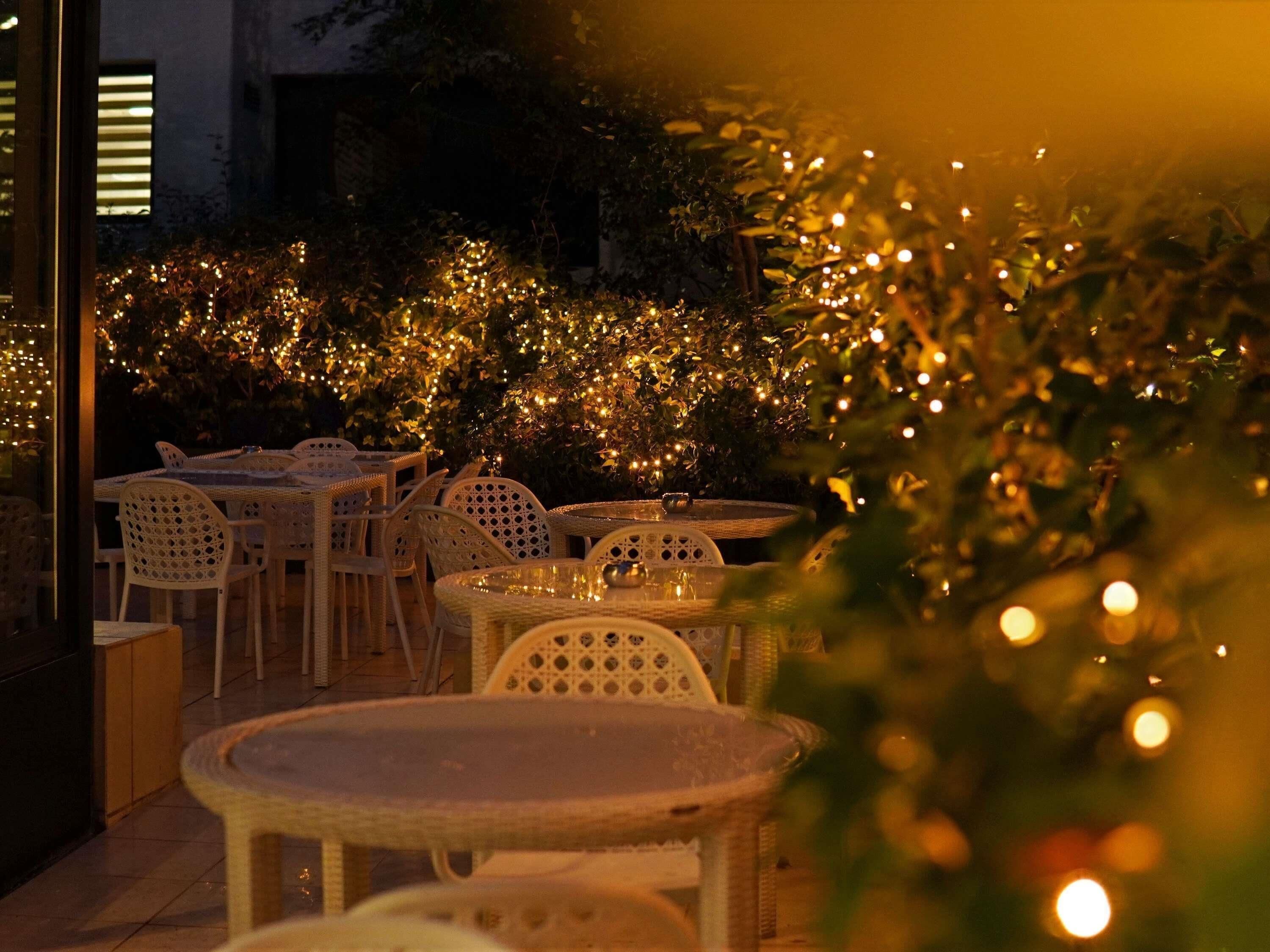 An outdoor dining area with wicker tables and chairs, surrounded by greenery and string lights.