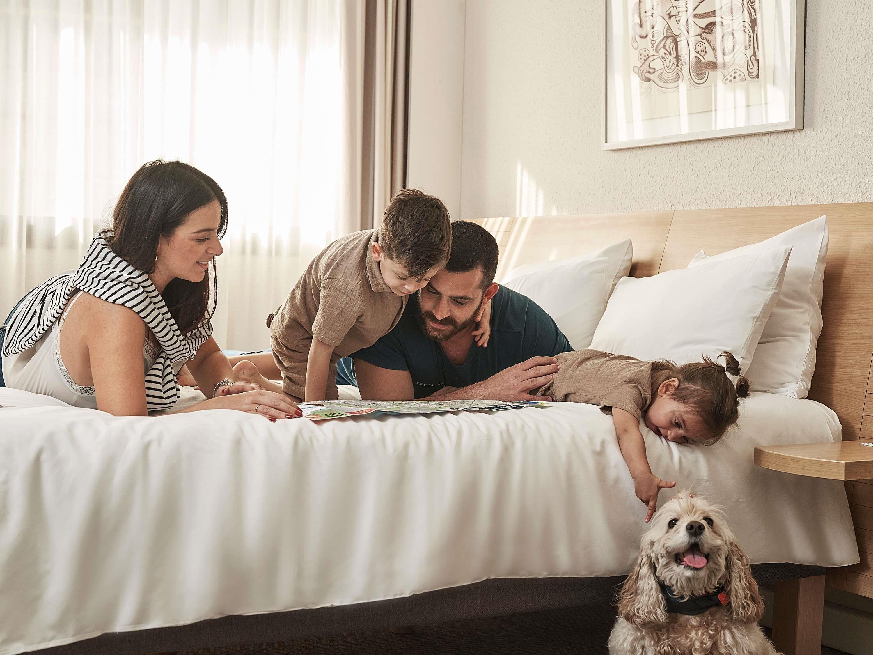 A family on a bed with a dog, in a room with a wooden headboard and a framed picture on the wall.