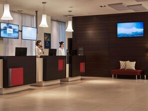 Een receptiegebied van een hotel met een modern design, met een receptiebalie, een televisiescherm en een zithoek met een rode bank.