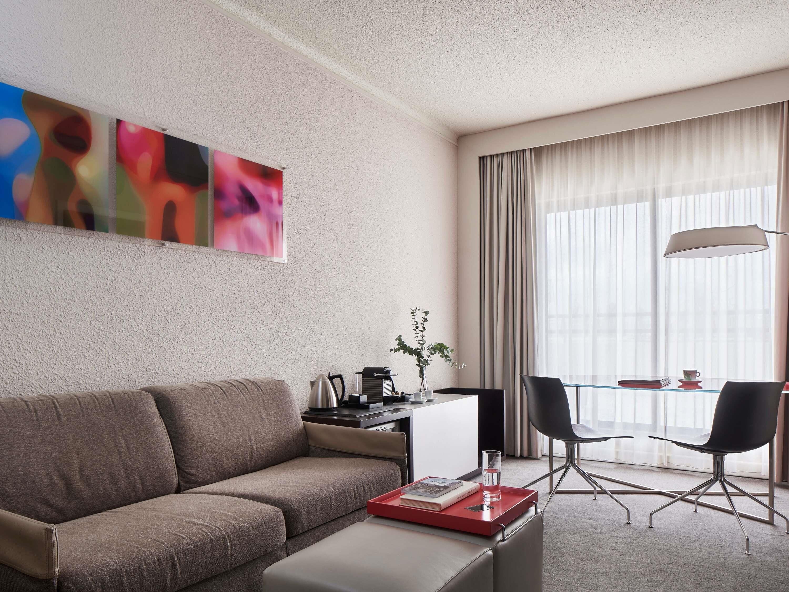 A modern hotel room with a sofa, a small table with a lamp, a chair, and a large window with curtains.