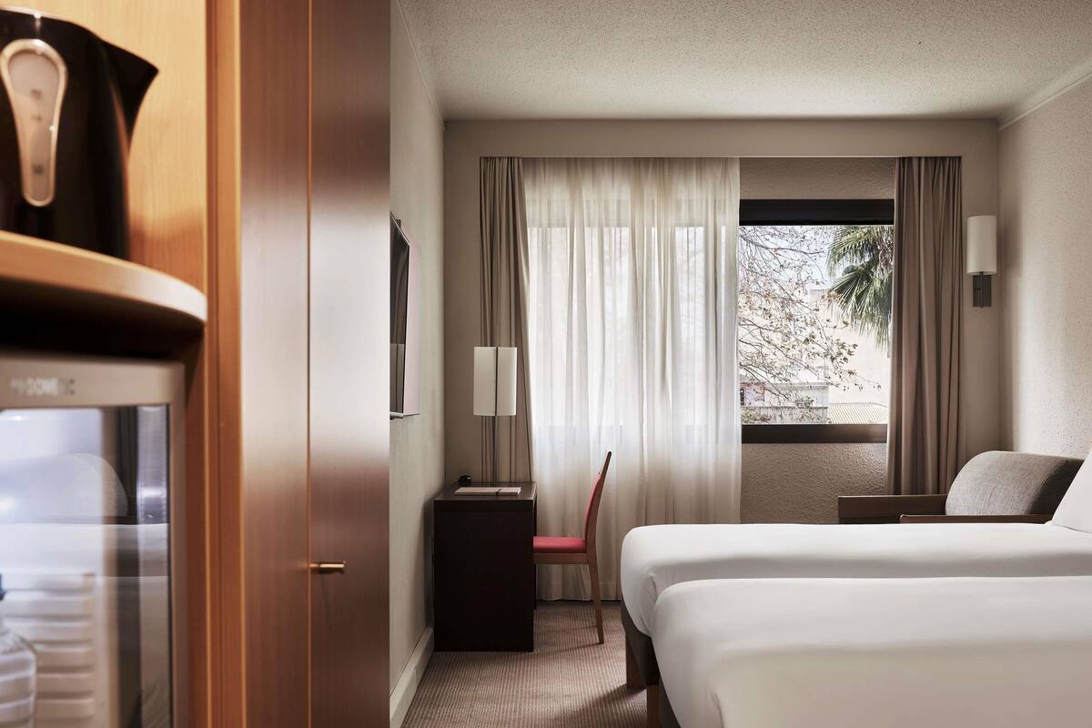 A hotel room with a bed, a desk, a chair, a lamp, and a window with curtains.