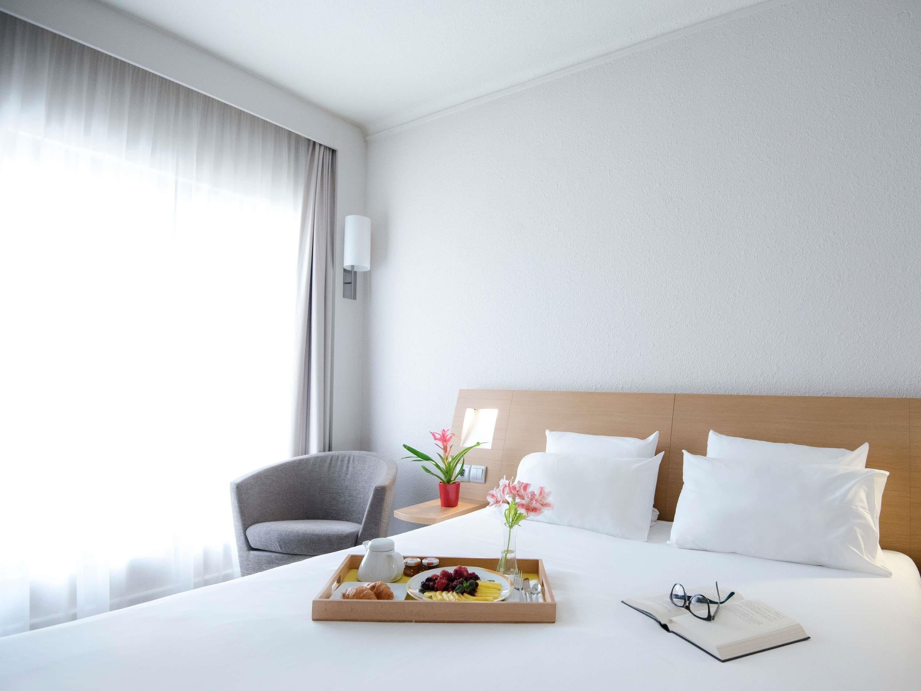 A hotel room with a bed, a bedside table with a tray of breakfast, a chair, a potted plant, and a window with curtains.