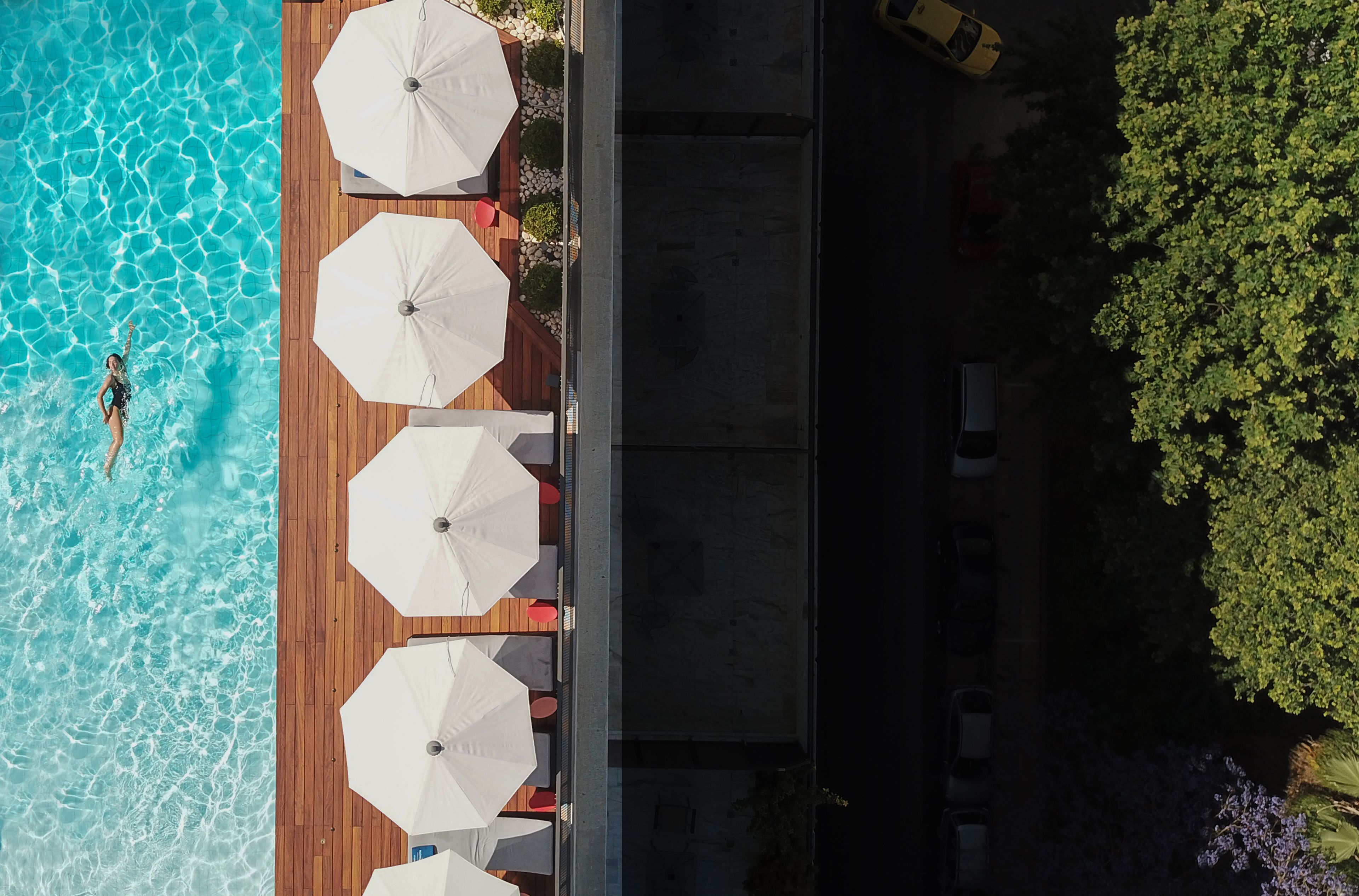 Vue aérienne d’un espace autour d’une piscine, avec une terrasse en bois, des parasols blancs et une personne qui nage.
