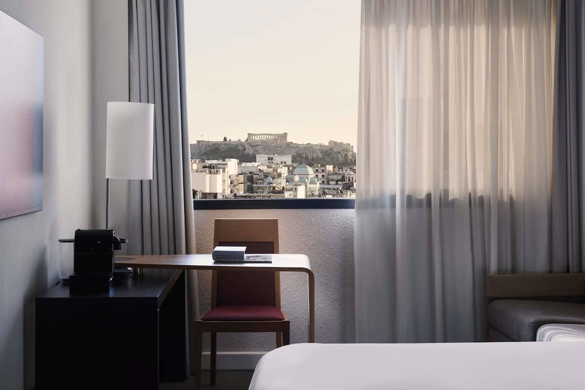 A hotel room with a bed, a desk with a lamp, a chair, and a view of a cityscape with historical buildings.