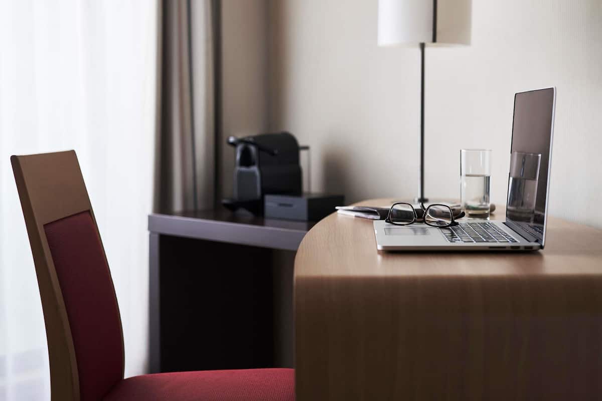 A hotel room with a desk, a laptop, a pair of glasses, a glass of water, and a red chair.