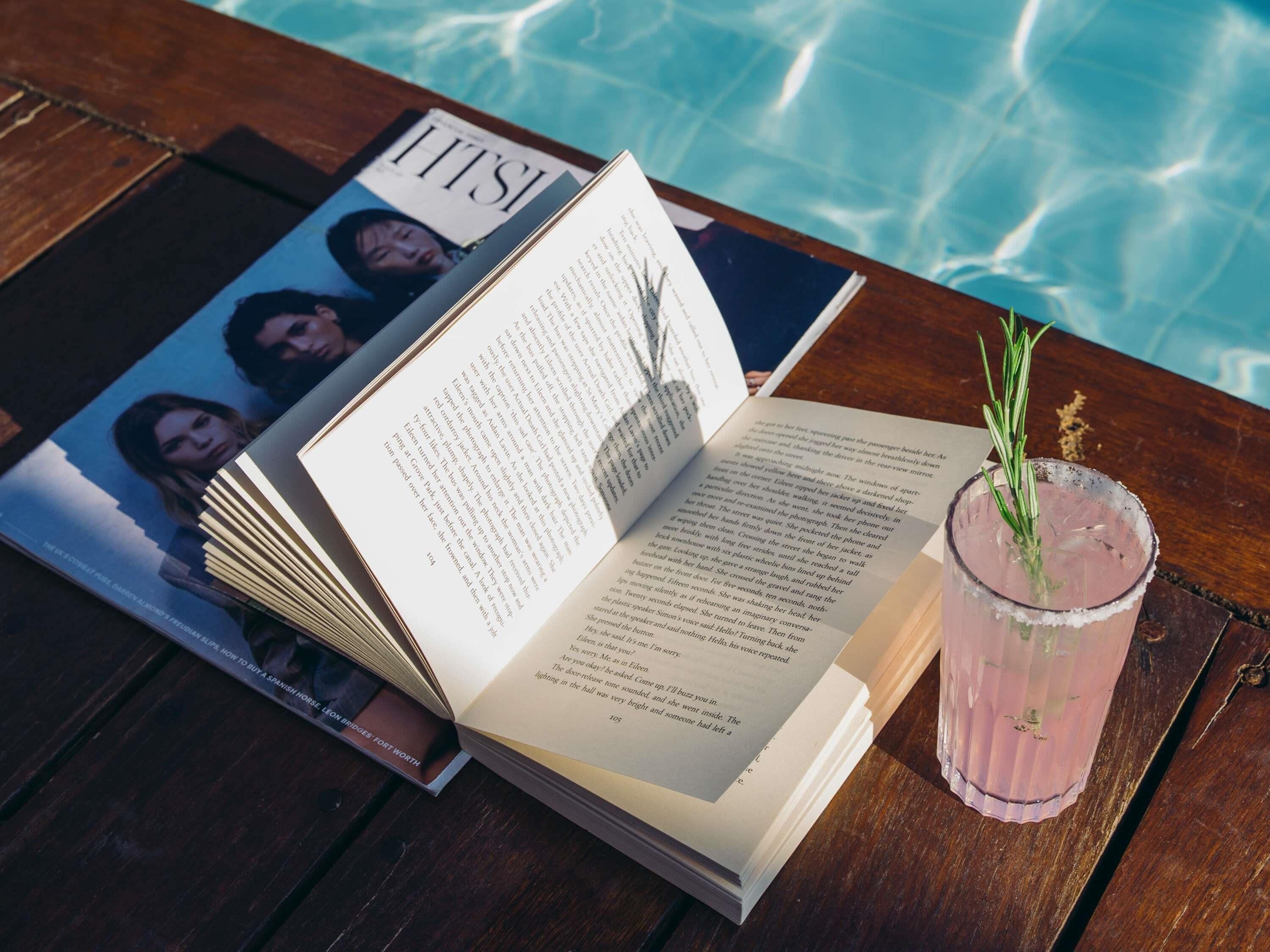 A book, a magazine, and a drink on a wooden table by a pool.
