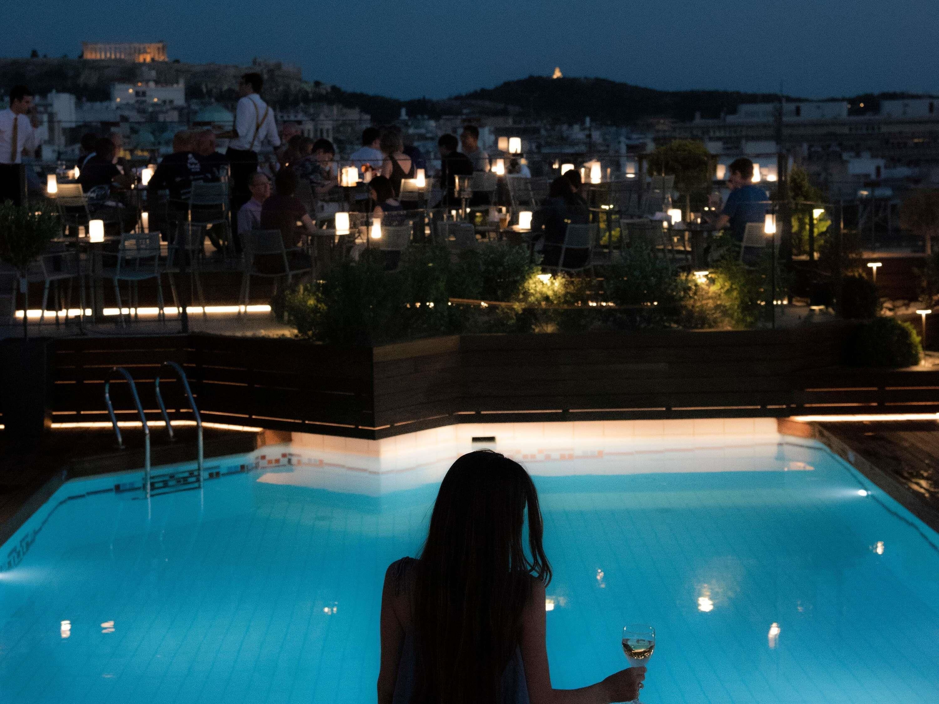 Zona de piscina con una persona sosteniendo un vaso, rodeada de sillas y mesas con iluminación.