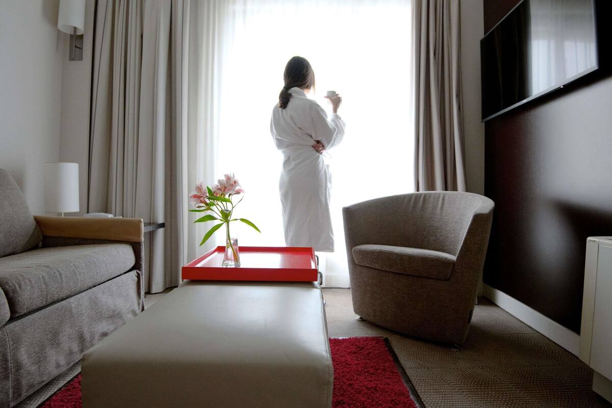 A modern hotel room with a sofa, armchair, a red tray with flowers, and a large window with curtains.