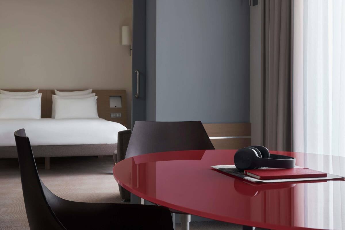 A hotel room with a bed, a desk with a chair, a lamp, and a window with curtains.
