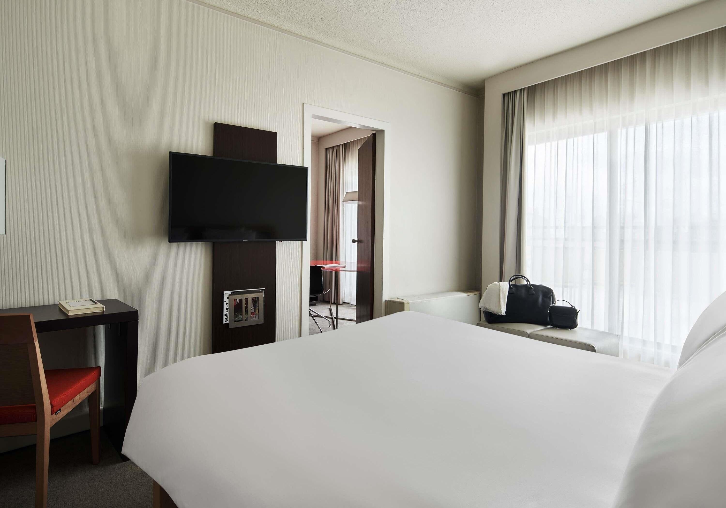Camera d'albergo con un letto grande, una scrivania con una sedia, una TV a schermo piatto fissata al muro, una finestra con tende leggere e un tavolino con una borsa.