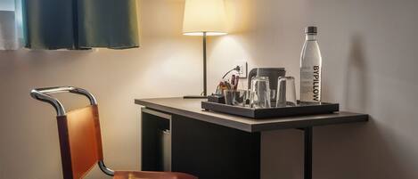 Habitación de hotel con escritorio, silla, lámpara, botella y cafetera.