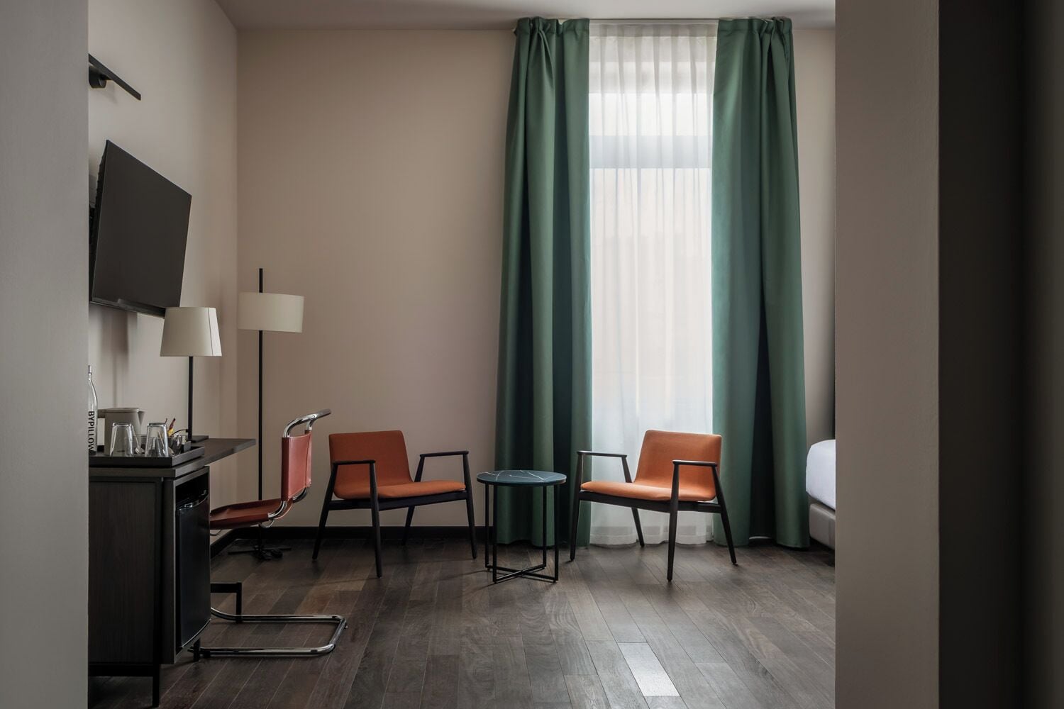 Habitación de hotel con televisión, dos sillas, una mesita, lámpara y ventana con cortinas.