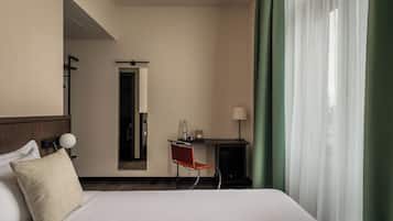 A hotel room with a bed, a desk, a chair, a lamp, and a view through sheer curtains.