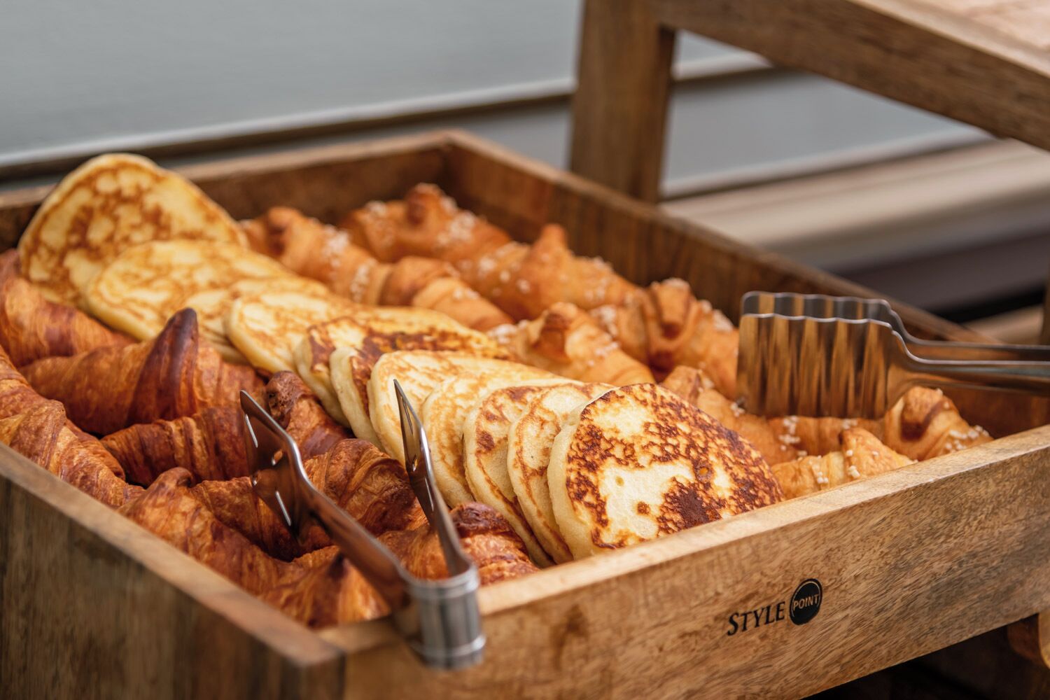 Una bandeja de madera con diversos pasteles, incluyendo croissants y tortitas, y dos pinzas metálicas.