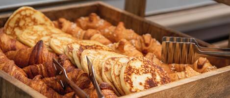 Una bandeja de madera con diversos pasteles, incluyendo croissants y tortitas, y dos pinzas metálicas.