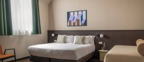 A hotel room with a large bed, a smaller bed, a chair, a bedside table with a bottle and glass, and a wall-mounted artwork.