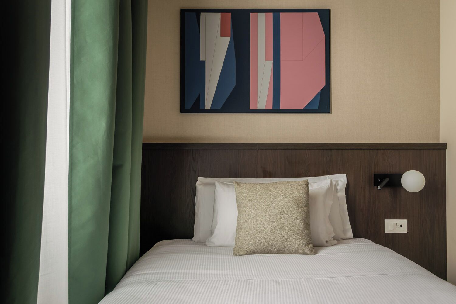 A hotel room with a bed, pillows, a wall-mounted light, and a piece of abstract art.