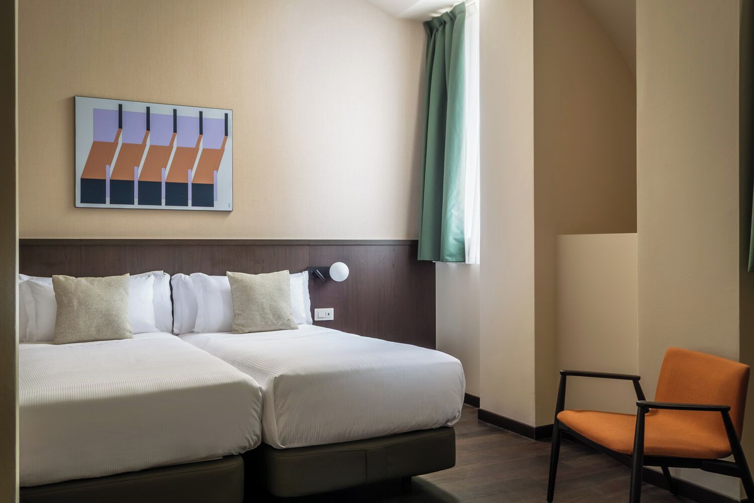A hotel room with a double bed, a chair, a wall-mounted light, and a painting.