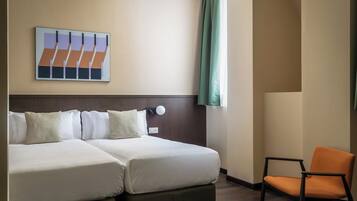 A hotel room with a double bed, a chair, a wall-mounted light, and a painting.