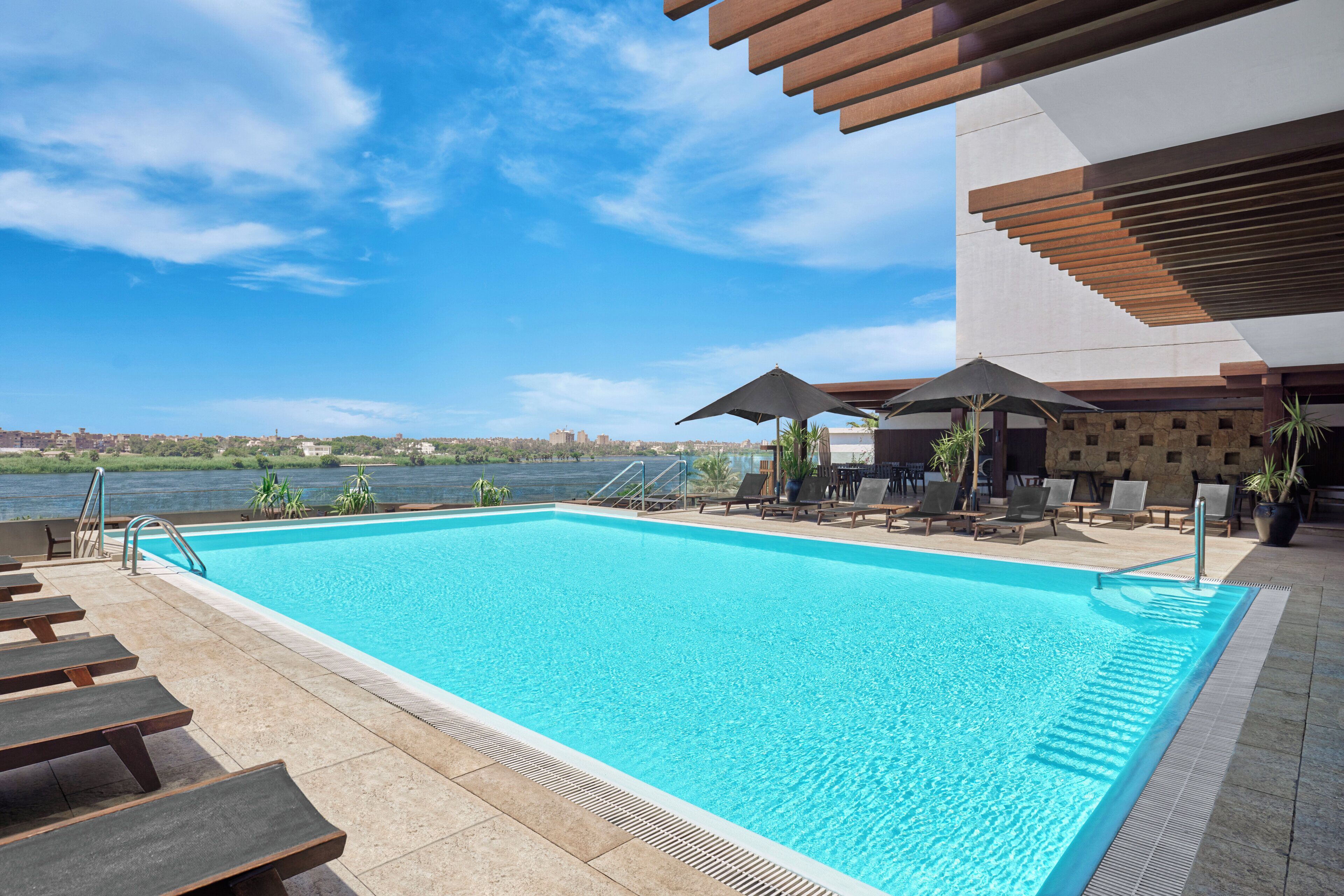 A swimming pool with lounge chairs and umbrellas, a view of the water, and a modern building structure.