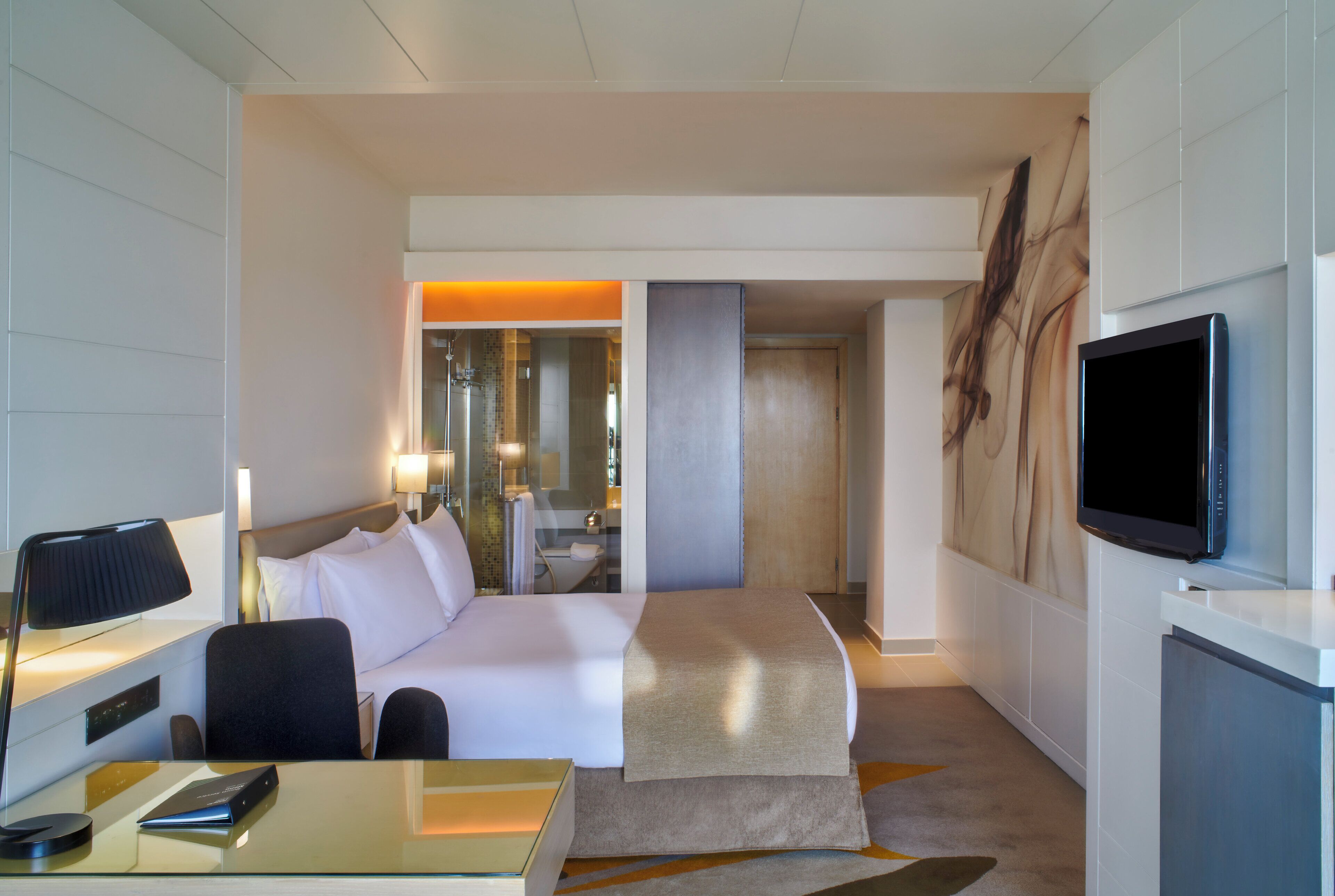 A hotel room with a bed, bedside table, lamp, TV, and a desk with a chair.