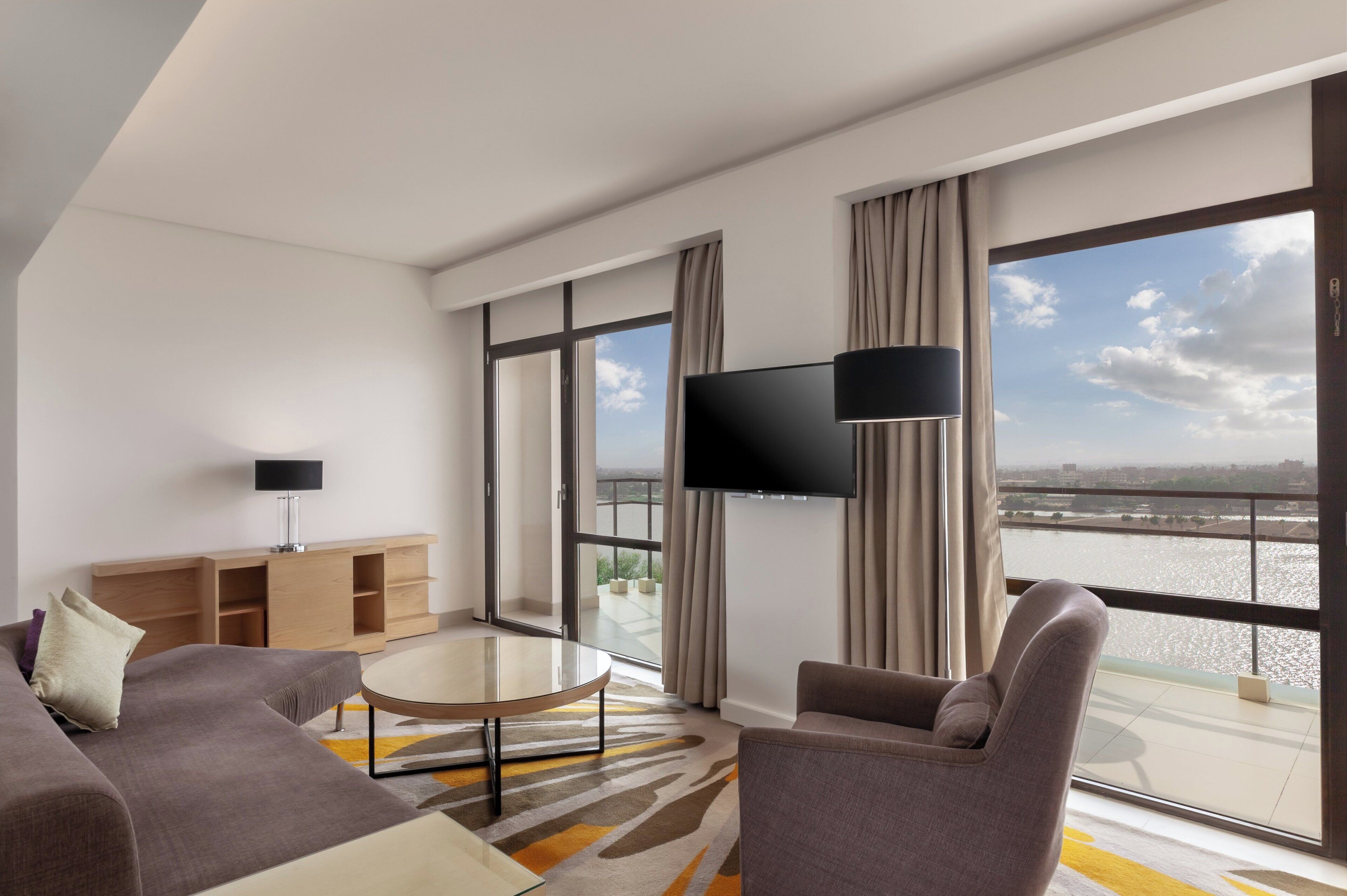 A modern hotel room with a large window, a flat-screen TV, a sofa, a coffee table, and a wooden side table.