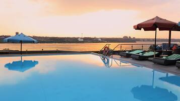 Una piscina con sillones de descanso y sombrillas, con vista a una masa de agua y un paisaje urbano al fondo.