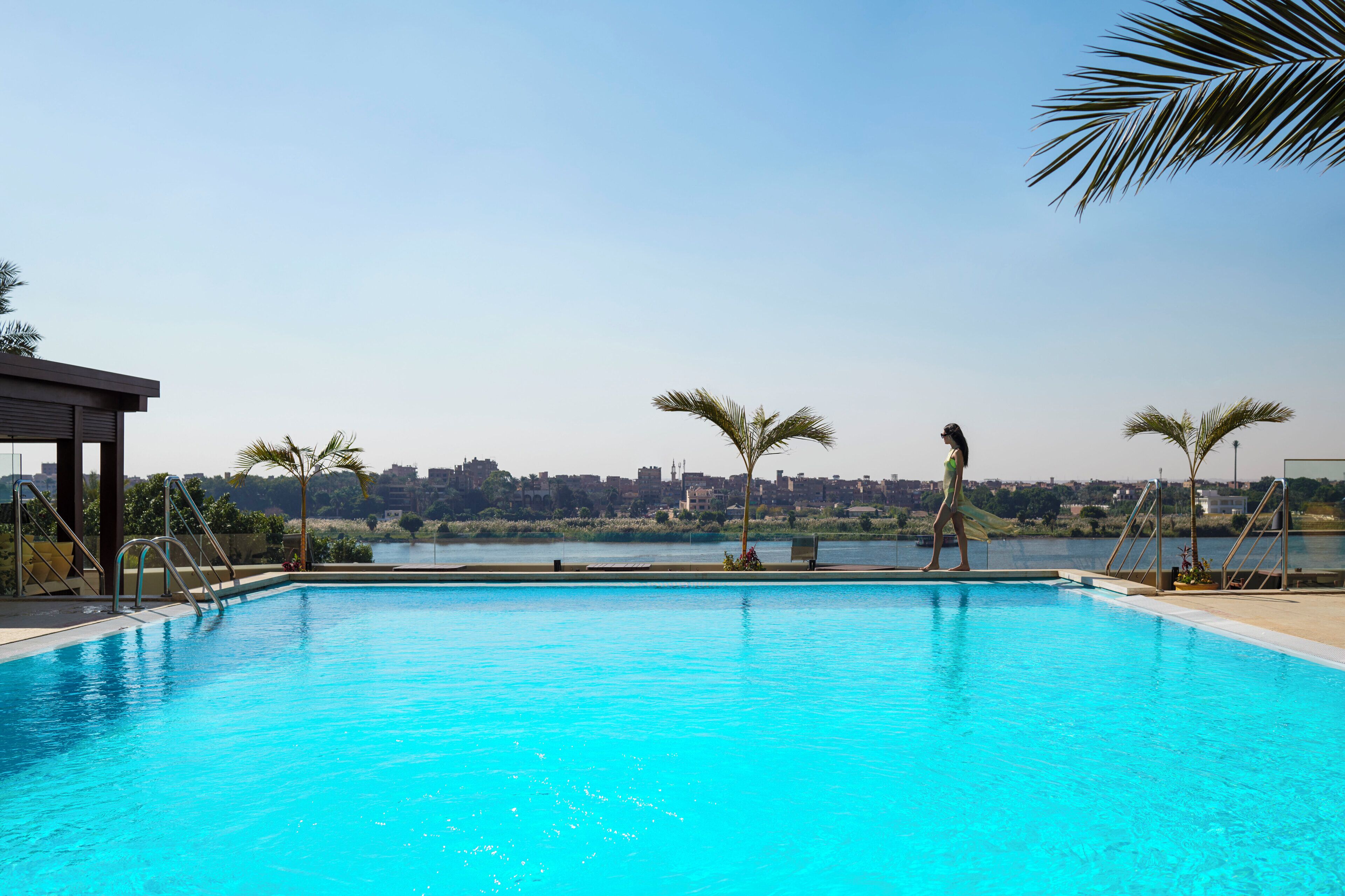 A swimming pool with clear blue water, surrounded by palm trees and a cityscape in the background.