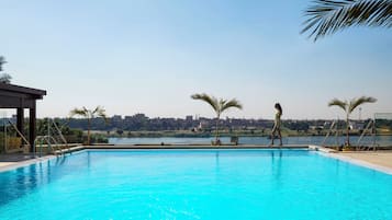 A swimming pool with clear blue water, surrounded by palm trees and a cityscape in the background.