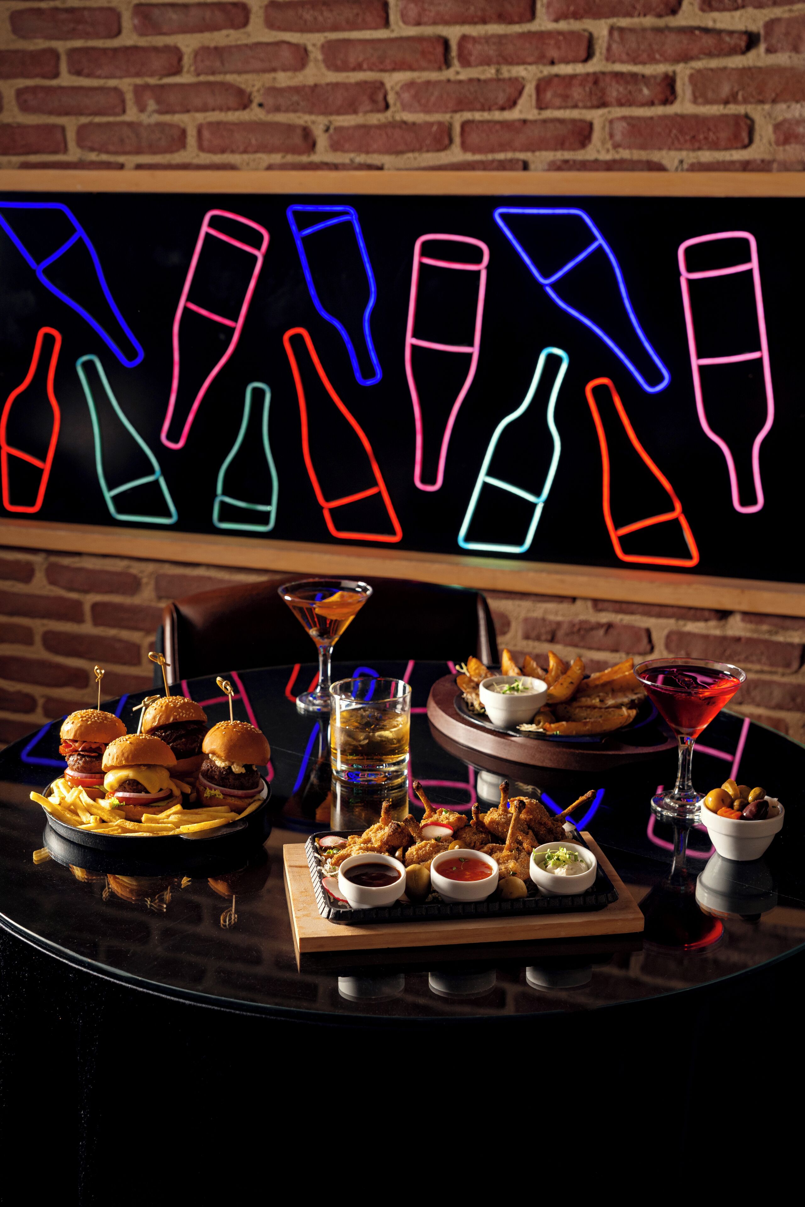 A table with food and drinks, a neon sign with bottle outlines, and a brick wall.