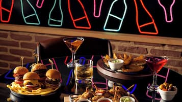 A table with food and drinks, a neon sign with bottle outlines, and a brick wall.